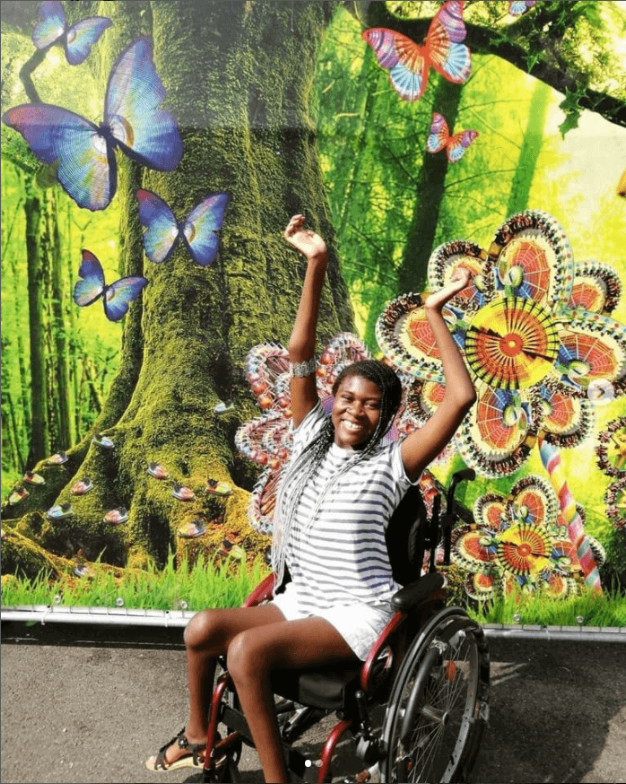 Simi in front of a colourful wall painted with image of trees and butterflies. She is smiling and holding her arms in the air.
