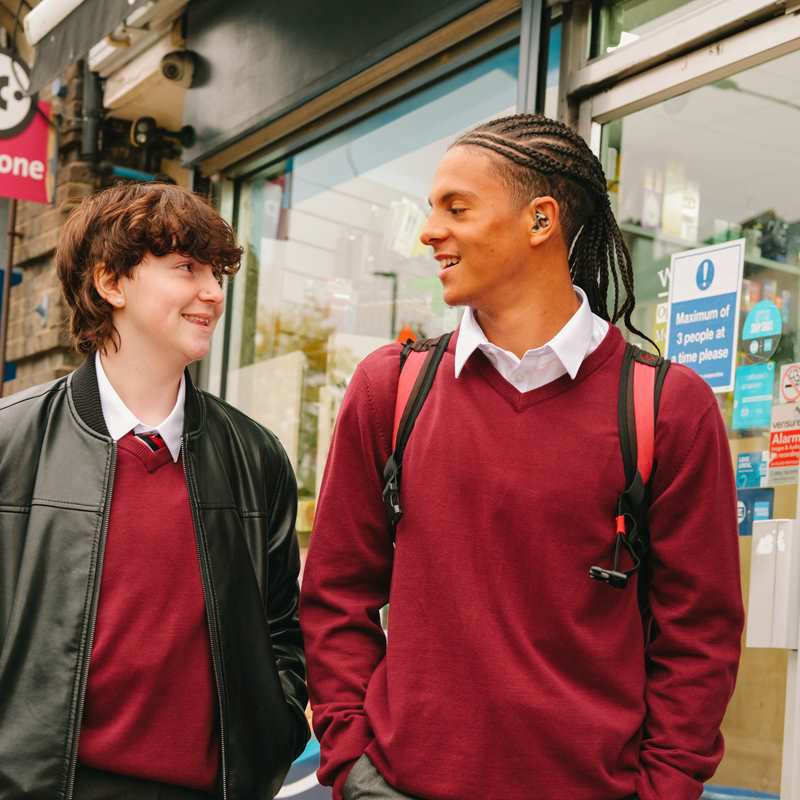 A Black teenage boy wearing a hearing aid speaking to a white non-binary teenager. They are walking on the street outside a shop. Both people are smiling.