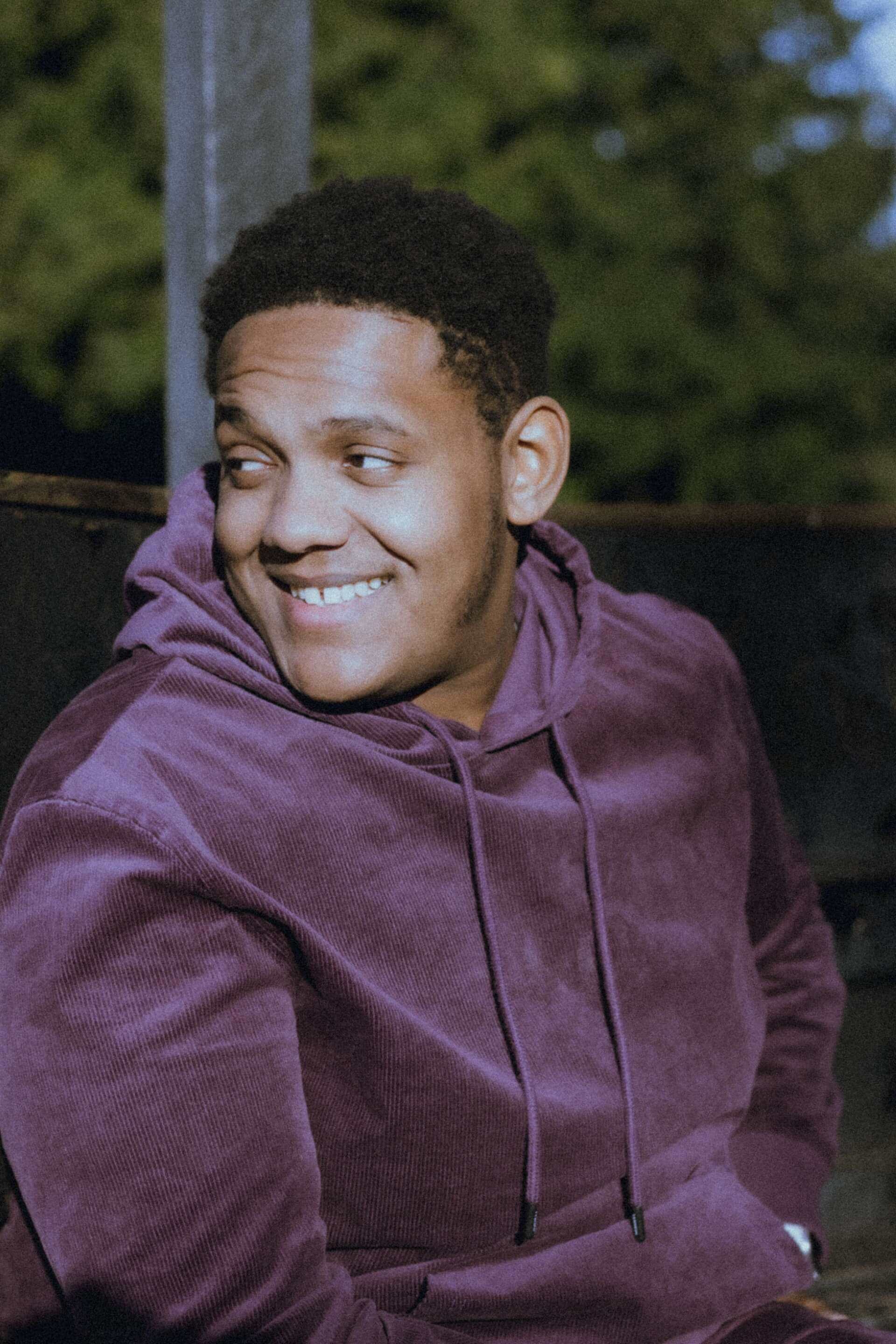 Josiah sitting in a park wearing a purple hoodie. He is looking over his right shoulder and smiling.