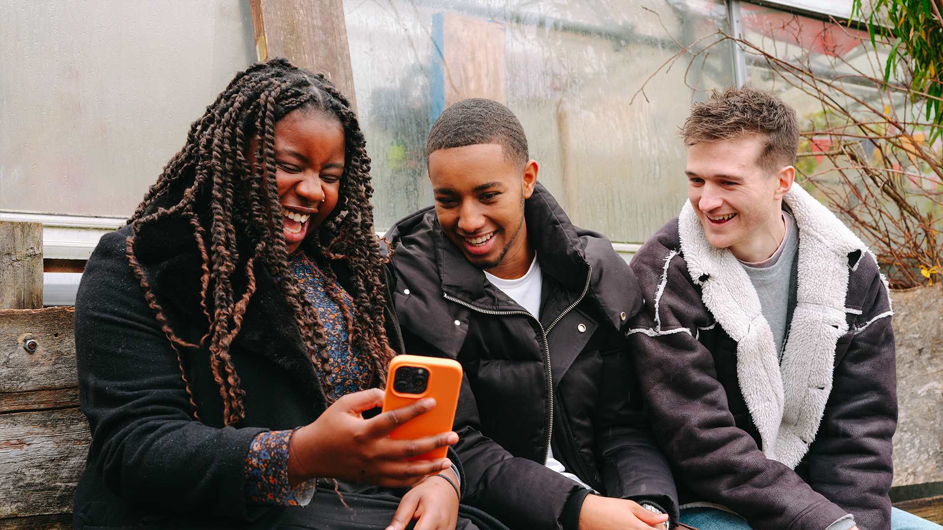 A young Black woman, young Black man and young white man, all sitting on a bench outside, looking at something on a phone and laughing.