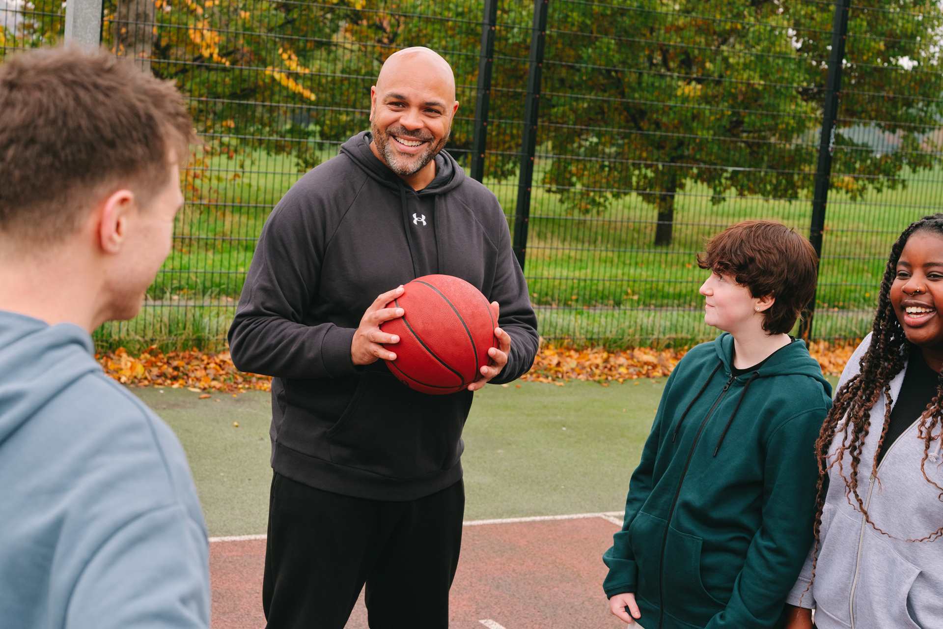 A group of young people playing basketball with an older Black man. The group of young people includes: one white young man, one white non-binary teenager and one Black young woman.