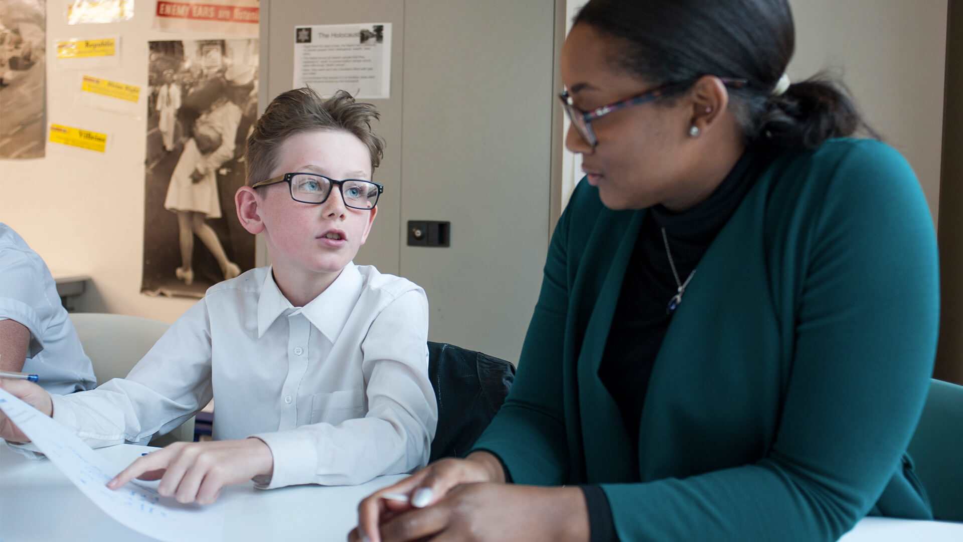 a boy wearing school uniform and glasses sits on his desk holding a paper asking help from his teacher sitting beside him in the classroom