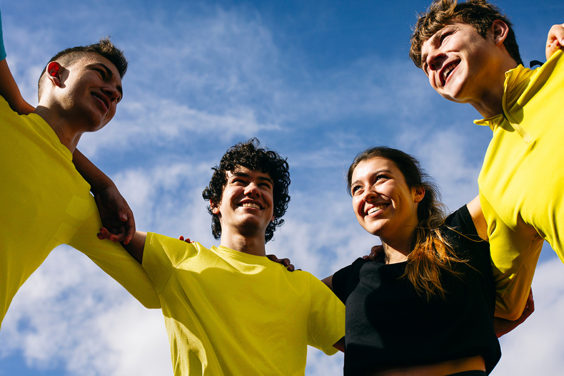 A group of young people with arms around each other in a circle, wearing yellow and smiling