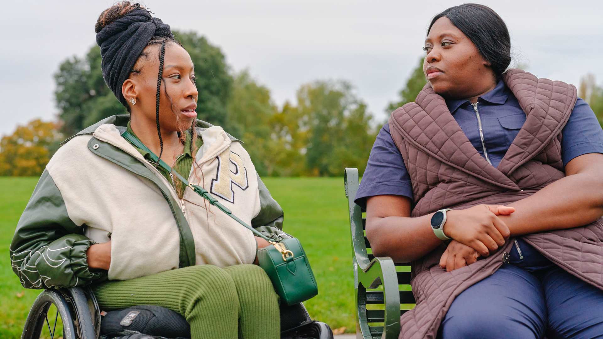 A young Black woman in a wheelchair and an older Black woman sitting on a bench in the park. They are talking about something serious.