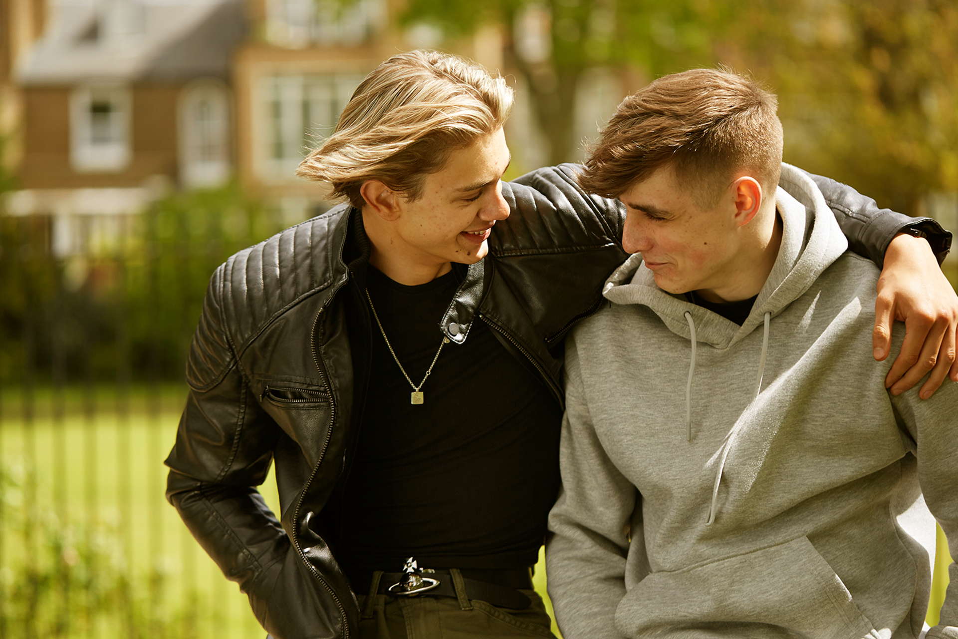 Two boys sitting in the park with their arms around each other, smiling and looking at each other.