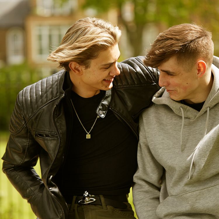 Two boys sitting in the park with their arms around each other, smiling and looking at each other.