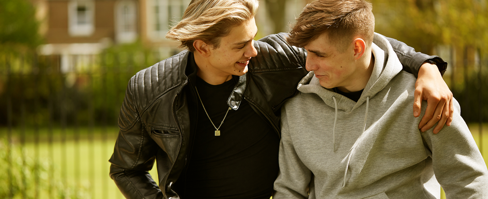 Two boys sitting in the park with their arms around each other, smiling and looking at each other.