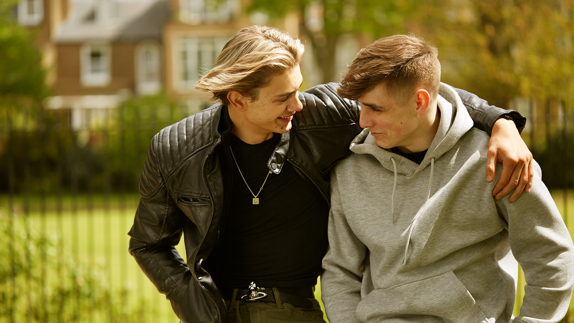 Two boys sitting in the park with their arms around each other, smiling and looking at each other.