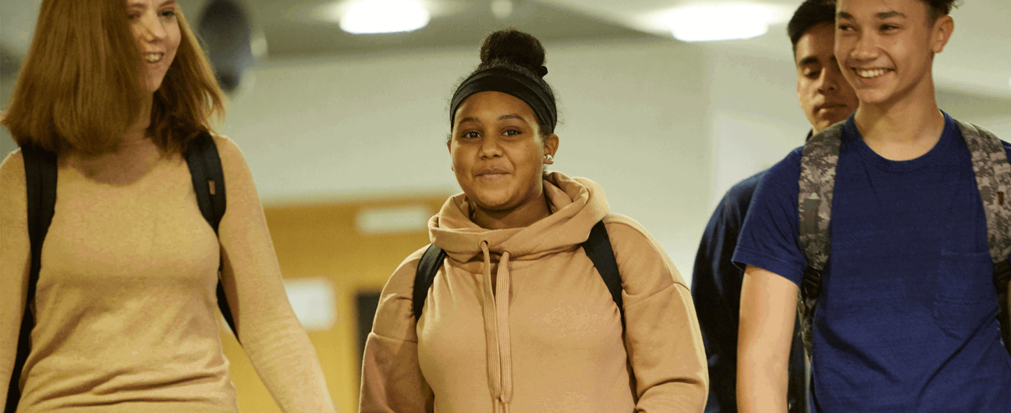Four students wearing backpacks and smiling while walking down a school hallway together.