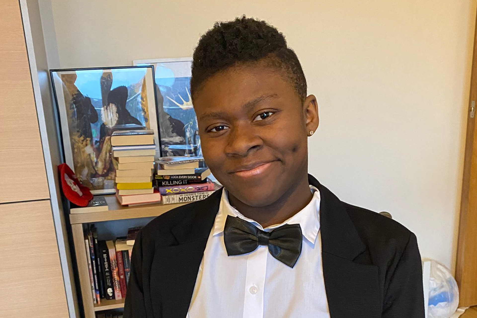 Simi is a Black young person and they are wearing a Black tuxedo with bow tie whilst sitting in their wheelchair and smiling.