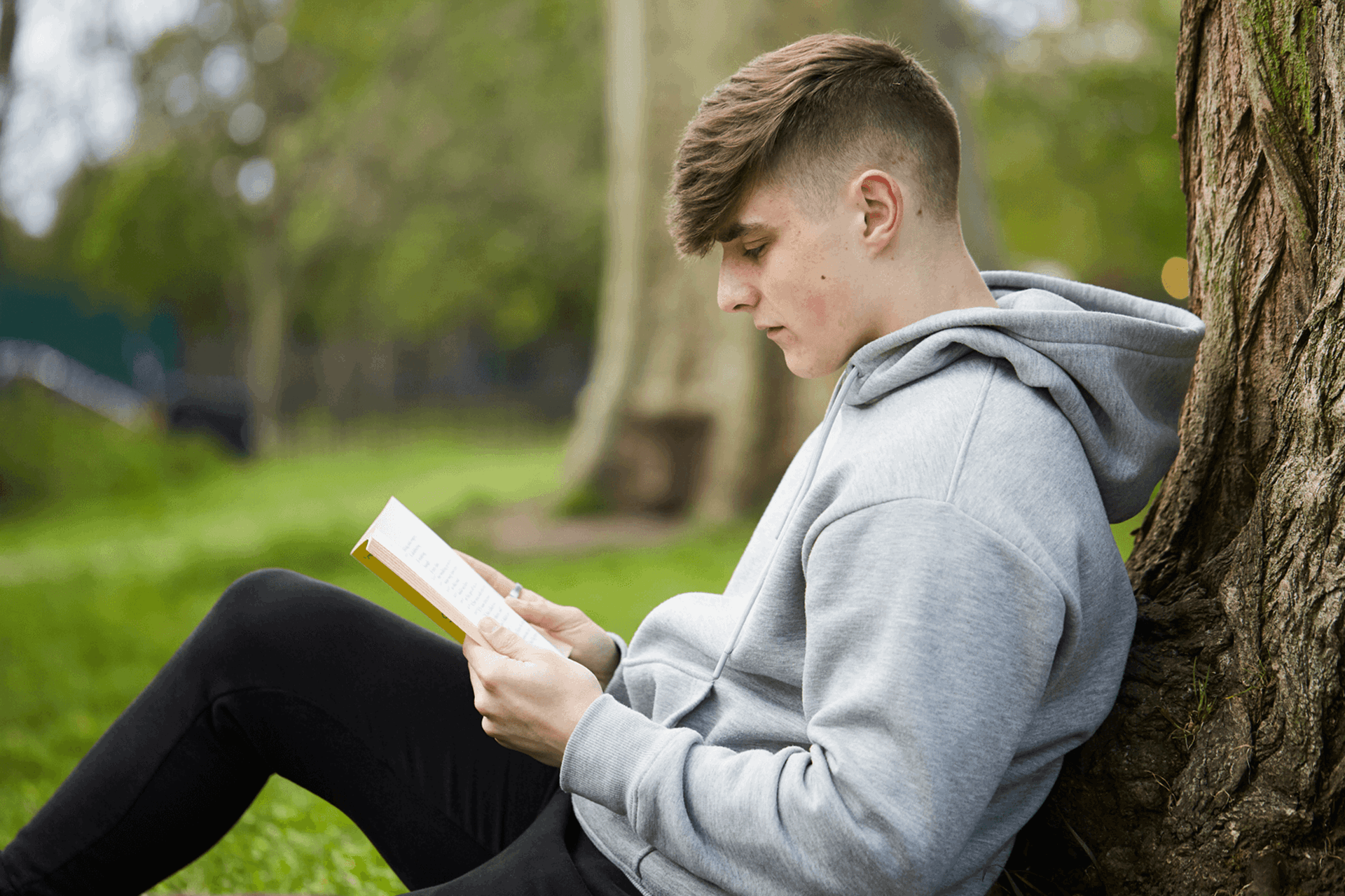 A young person sits against a tree in the park while reading a book.