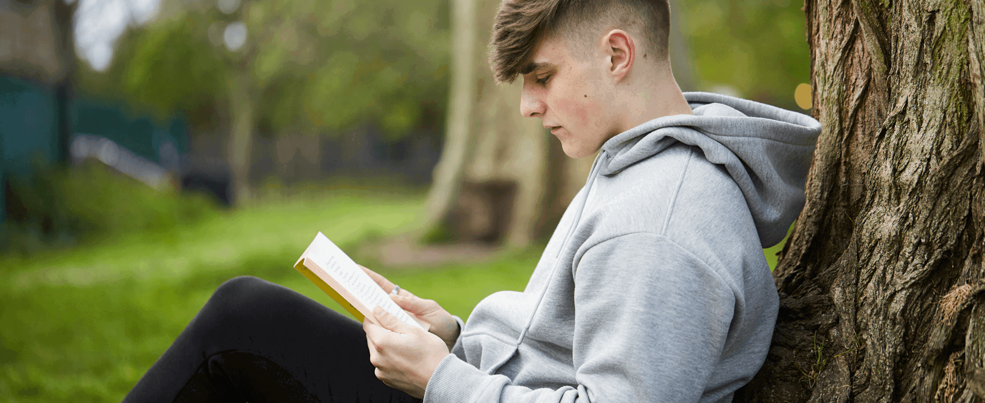 A young person sits against a tree in the park while reading a book.