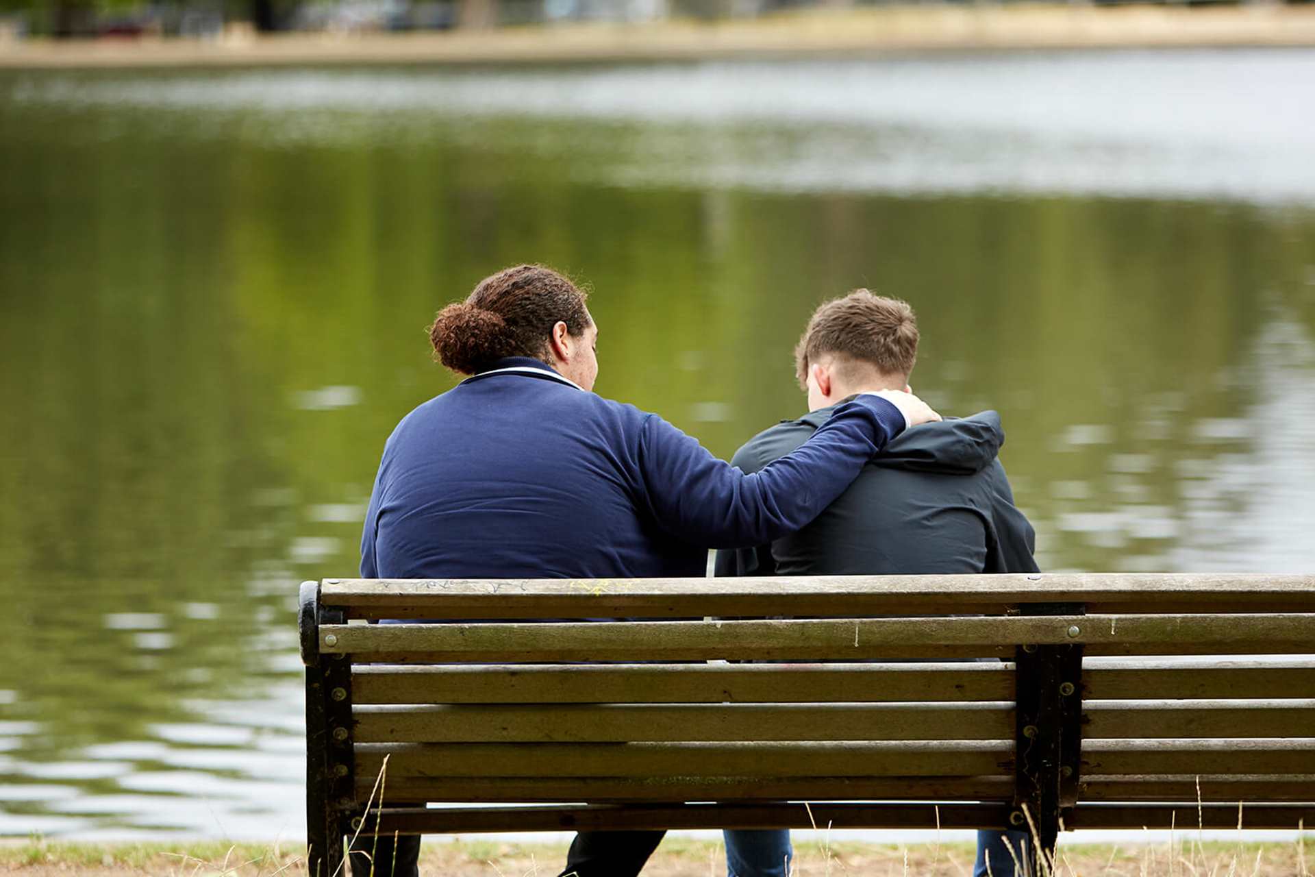 Two young men sit on a park bench overlooking a lake. One has his arm on the other's shoulder to comfort him.