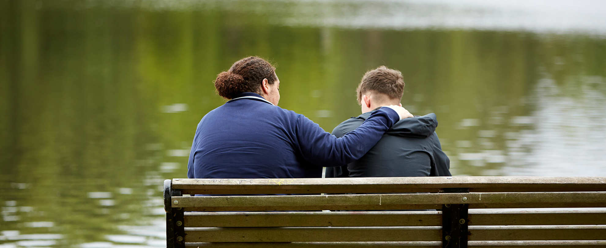 Two young men sit on a park bench overlooking a lake. One has his arm on the other's shoulder to comfort him.