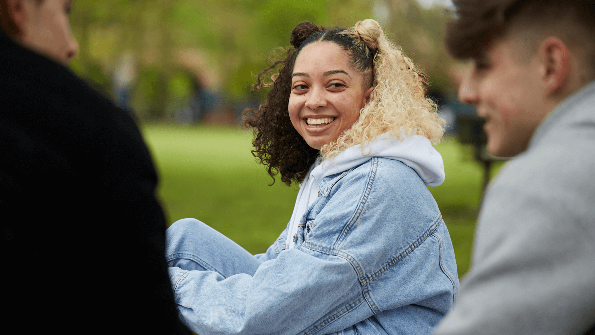 a-young-woman-wearing-a-blue-denim-jacket-and-with-curly-hair-smiling-to-two-young-man-talking-and-smiling-at-each-other-while-in-the-park