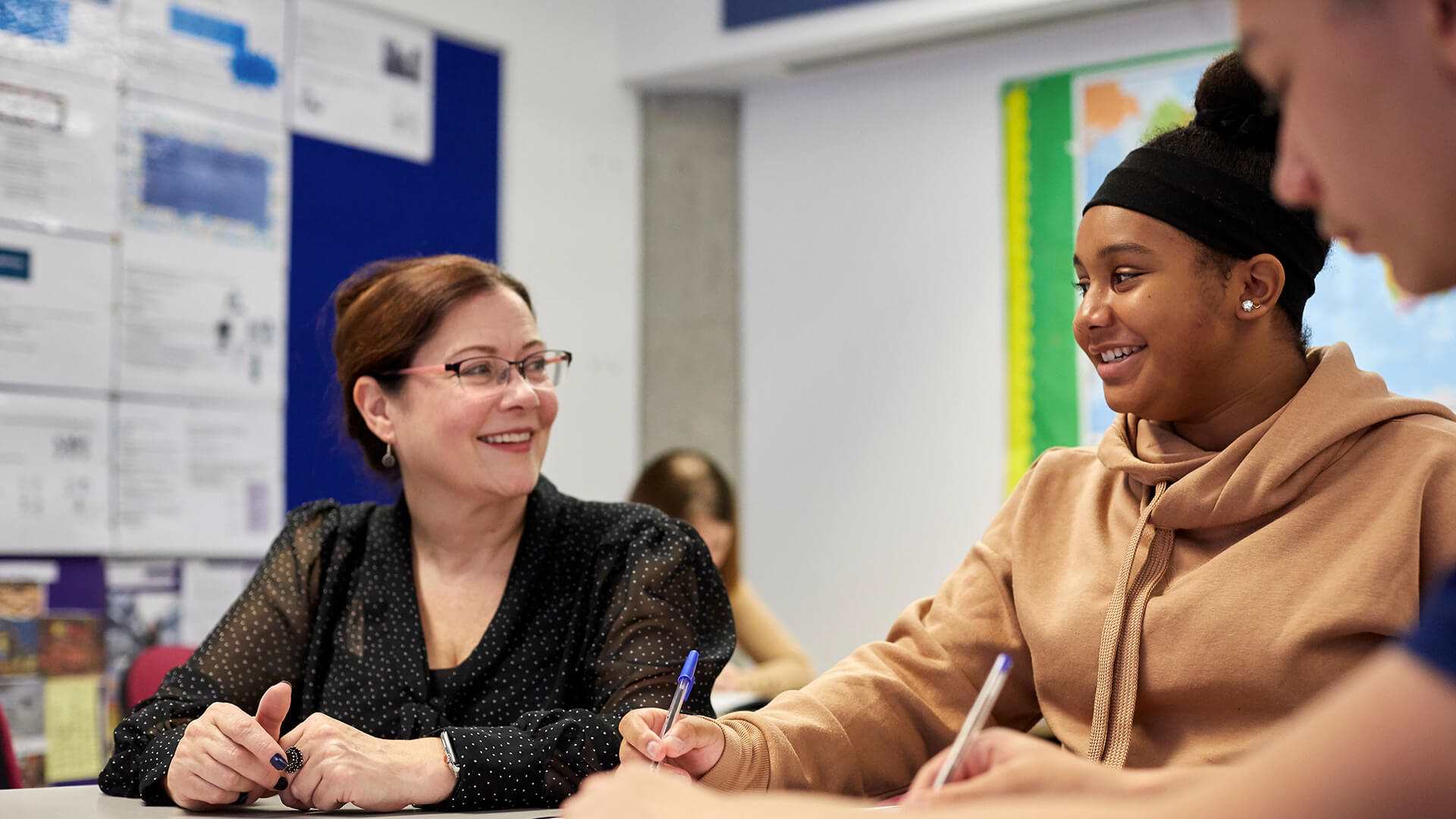 a teacher is smiling while she sits beside a student and help her working on an activity in class