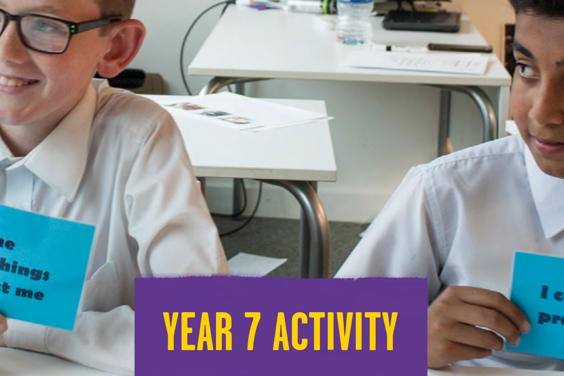 Two students sit next to each at a desk in a classroom holding blue cards with different statements on. In the middle of the students, there's a purple square with yellow writing. It says 'year 7 activity'.