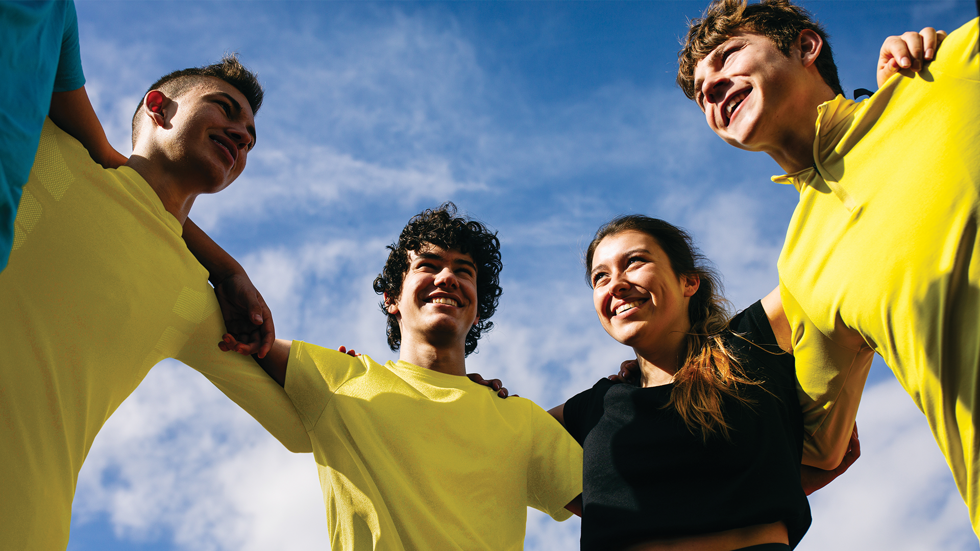 Four young people outdoors in yellow tshirts. They have their arms around each other and are all smiling. The camera is pointing up towards them to see the blue sunny sky and clouds in the background.