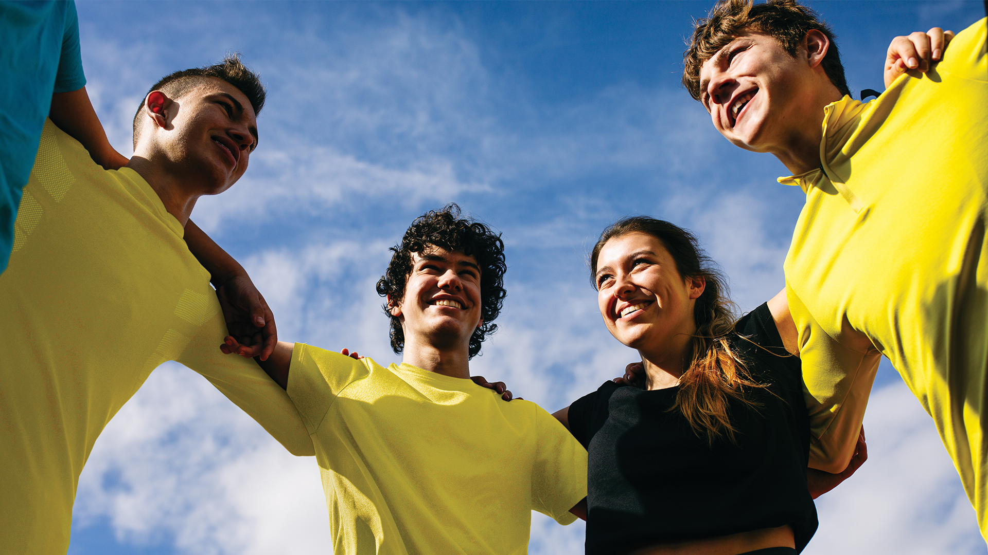 Four young people outdoors in yellow tshirts. They have their arms around each other and are all smiling. The camera is pointing up towards them to see the blue sunny sky and clouds in the background.