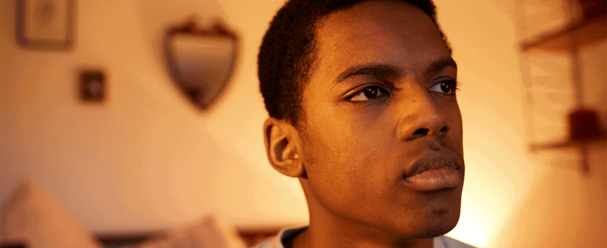 A boy sitting alone on the end of his bed and appearing deep in thought.