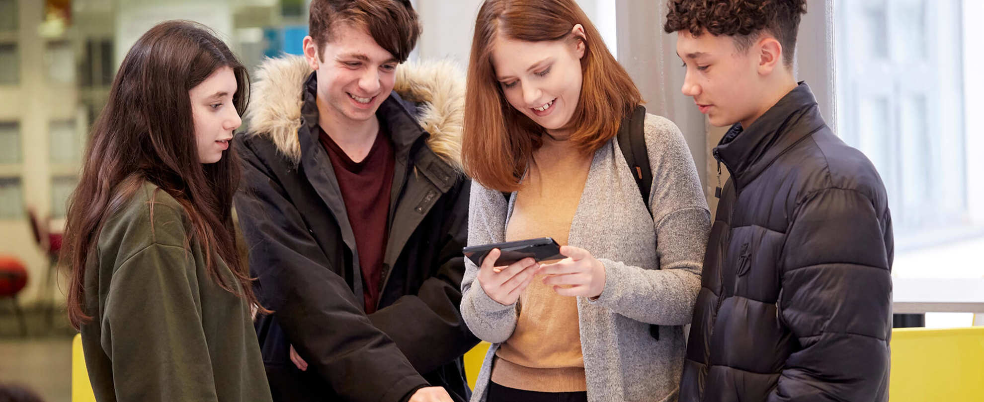 Four young people huddle round together, smiling and laughing, looking at a phone that the person in the middle is holding. They stand inside a campus building.