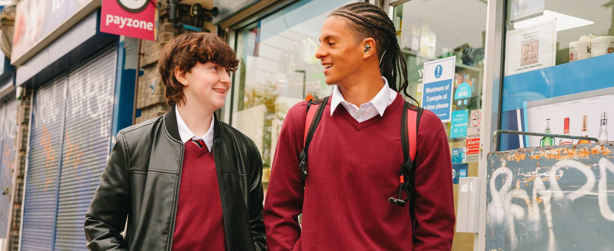 A Black teenage boy wearing a hearing aid speaking to a white non-binary teenager. They are walking on the street outside a shop. Both people are smiling.