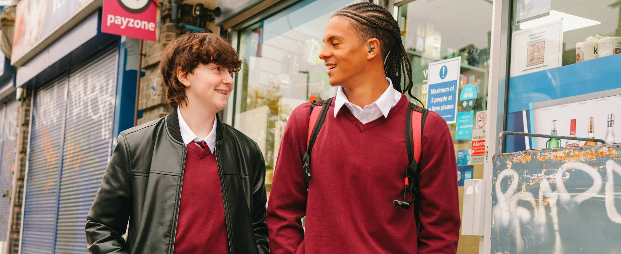 A Black teenage boy wearing a hearing aid speaking to a white non-binary teenager. They are walking on the street outside a shop. Both people are smiling.