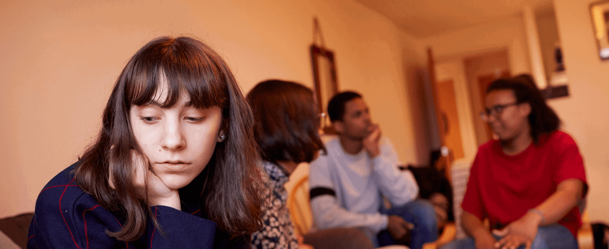 A girl sitting in a living room with her friends. She is looking away as they talk behind her and seems left out of the conversation.