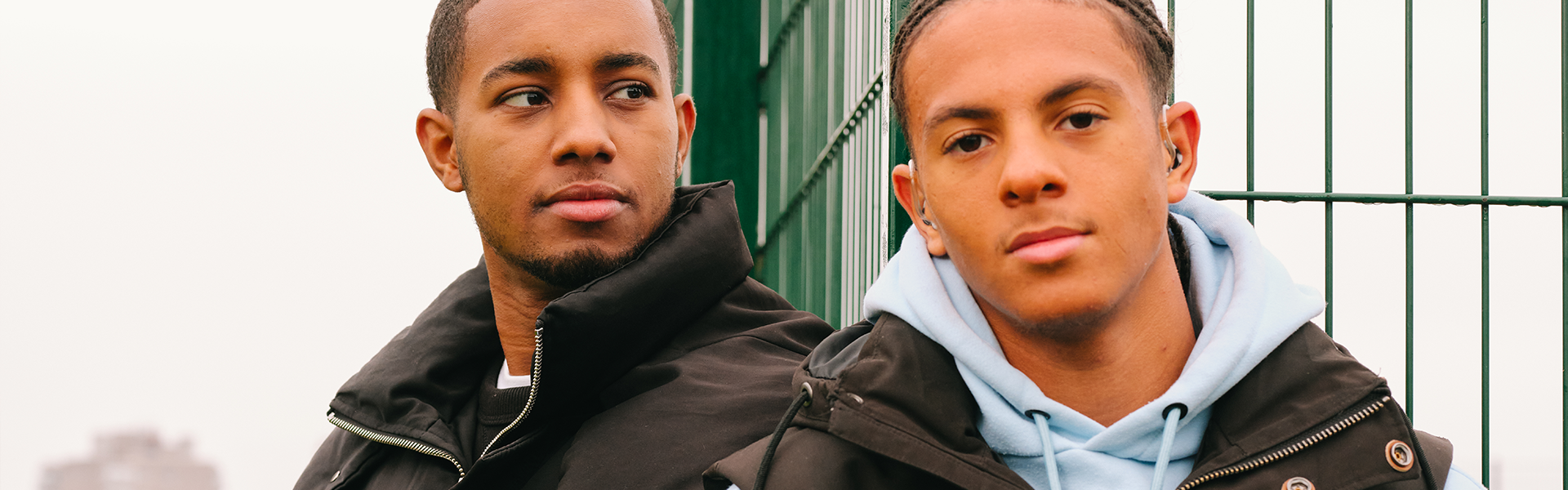 A young Black man sitting in the park with a Black teenage boy wearing a hearing aid. They are both looking very serious.