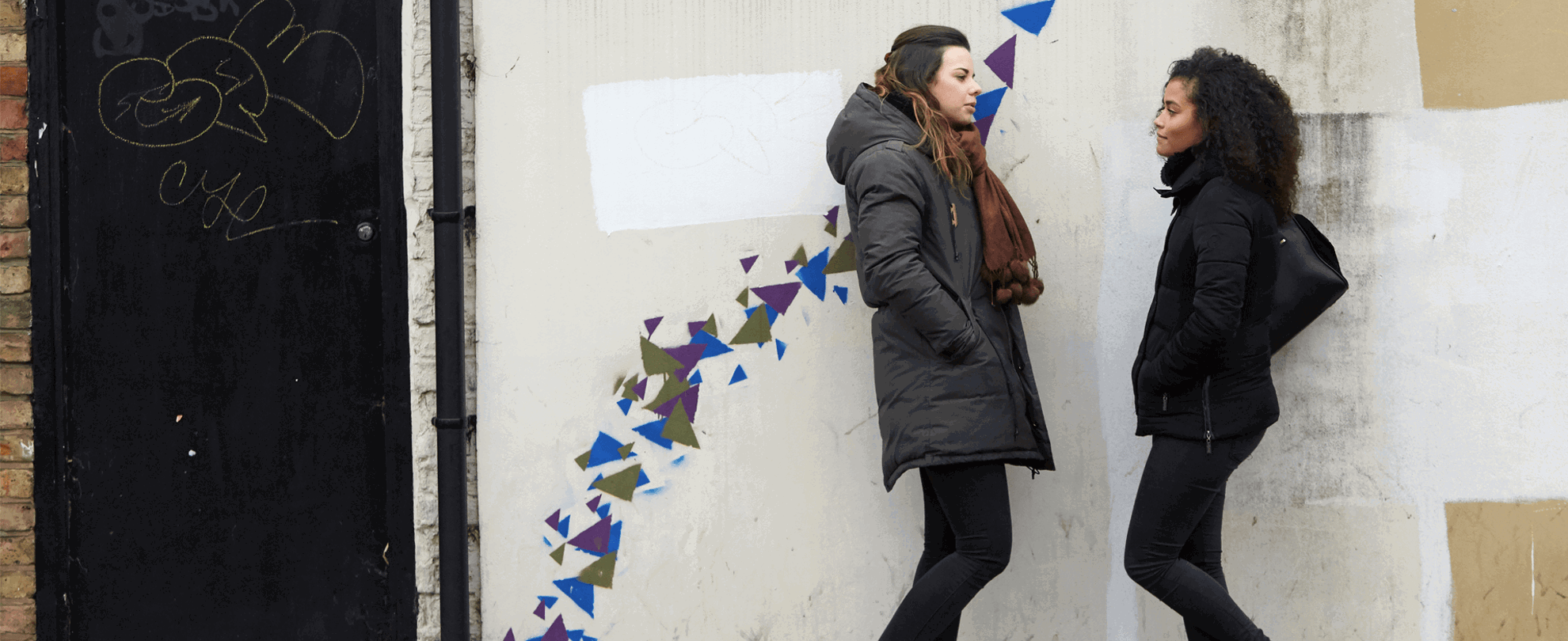 Two girls wearing black jackets are talking while standing against a wall decorated with blue, purple and green graffiti patterns.