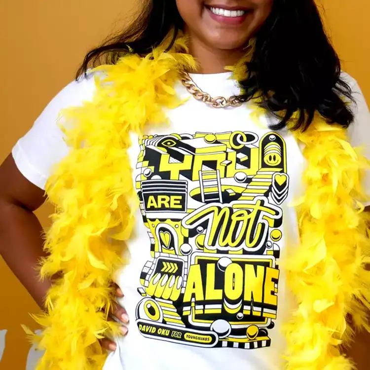 A close up of a woman smiling wearing a yellow feather bower, gold chain and our David Oku's bespoke 'You are not alone' graphic Hello Yellow T-shirt.