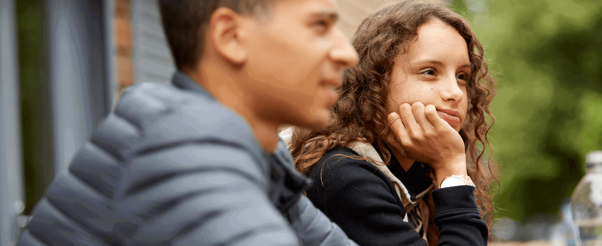 A girl with curly hair sits with her hand on her chin thinking, while a boy sits beside her wearing a grey jacket.