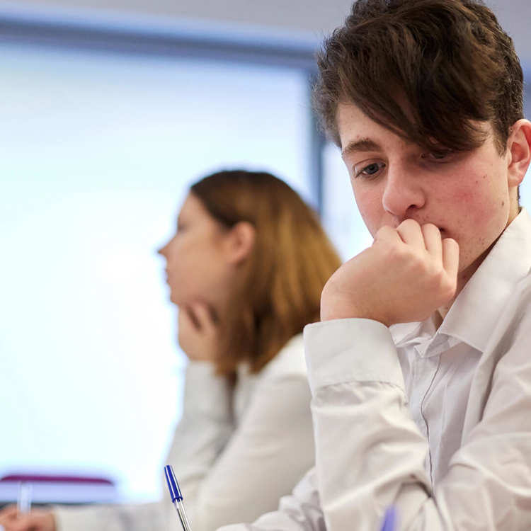 A student wearing uniform sits on a desk lost in a thought with their hand over their mouth, they sit next to another student who is focused on the lesson.