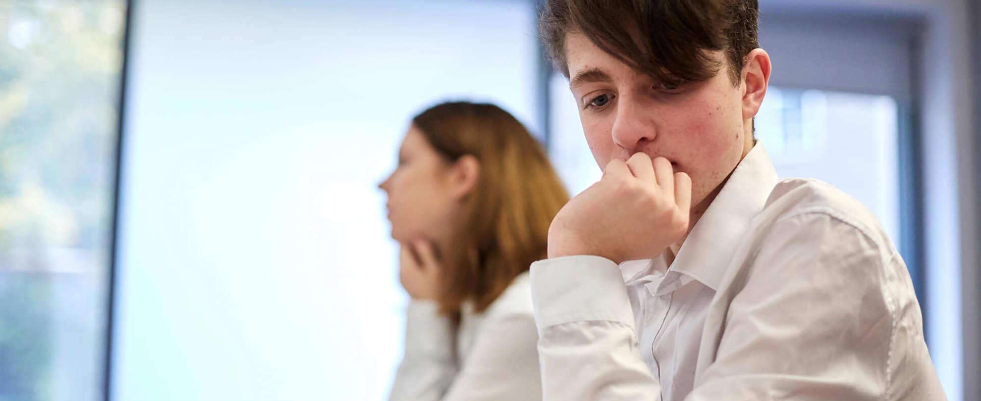 A student wearing uniform sits on a desk lost in a thought with their hand over their mouth, they sit next to another student who is focused on the lesson.
