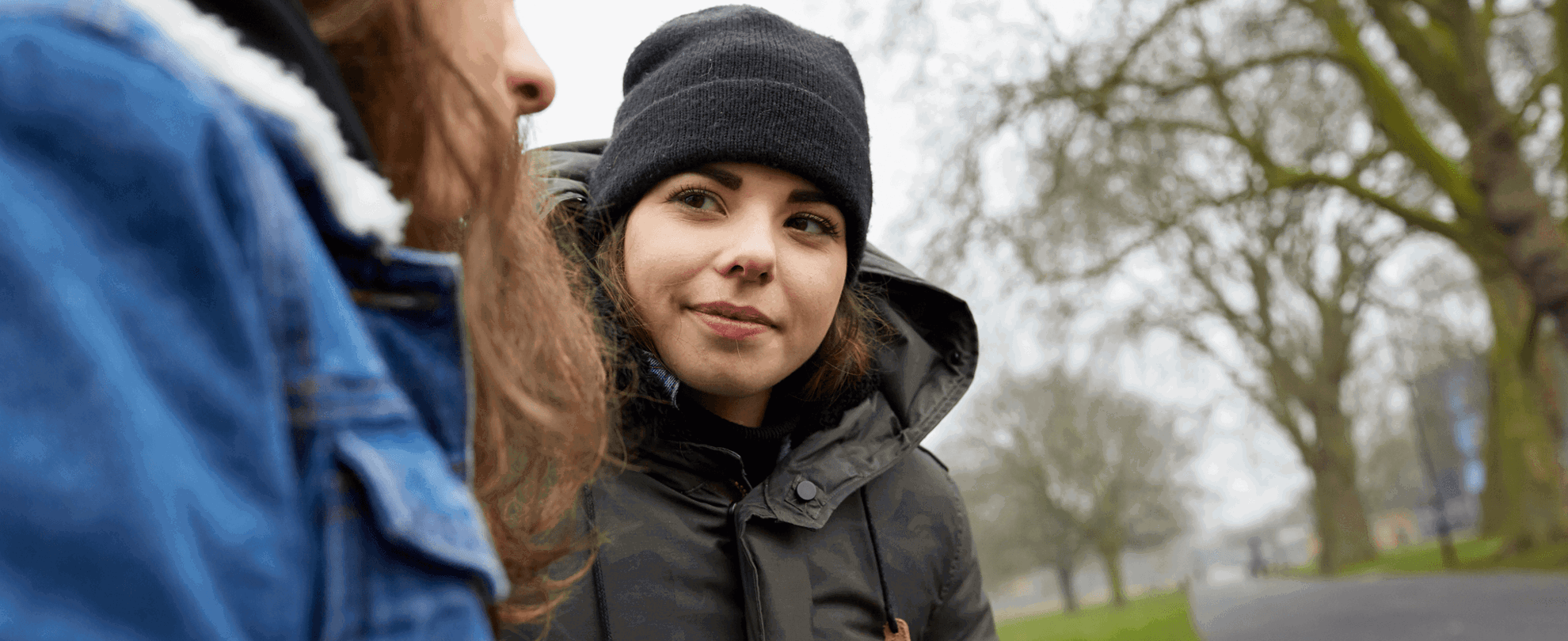 A girl in her winter coat is looking and smiling at her friend as they are talking while sitting on a park bench.