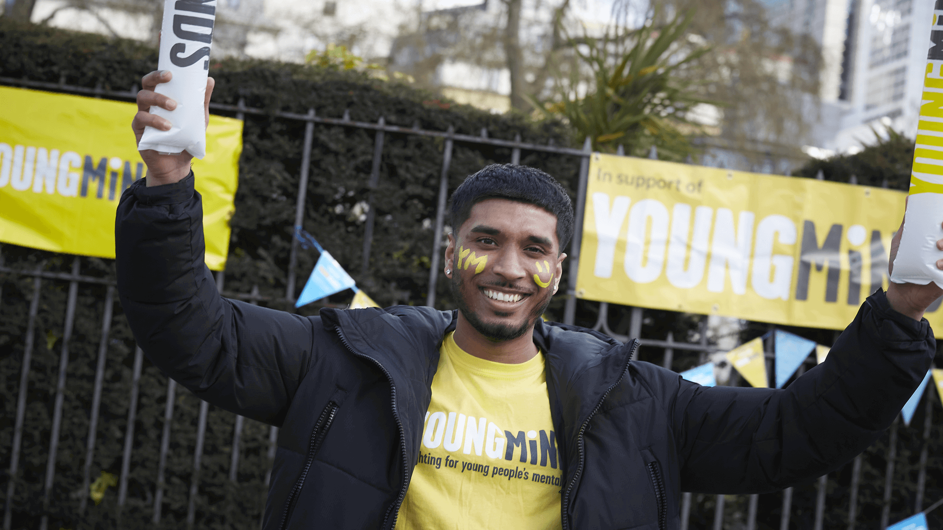 Volunteer in YoungMinds top, yellow face paint and black coat smiles in front of bunting and banners, with arms up holding cheer sticks.