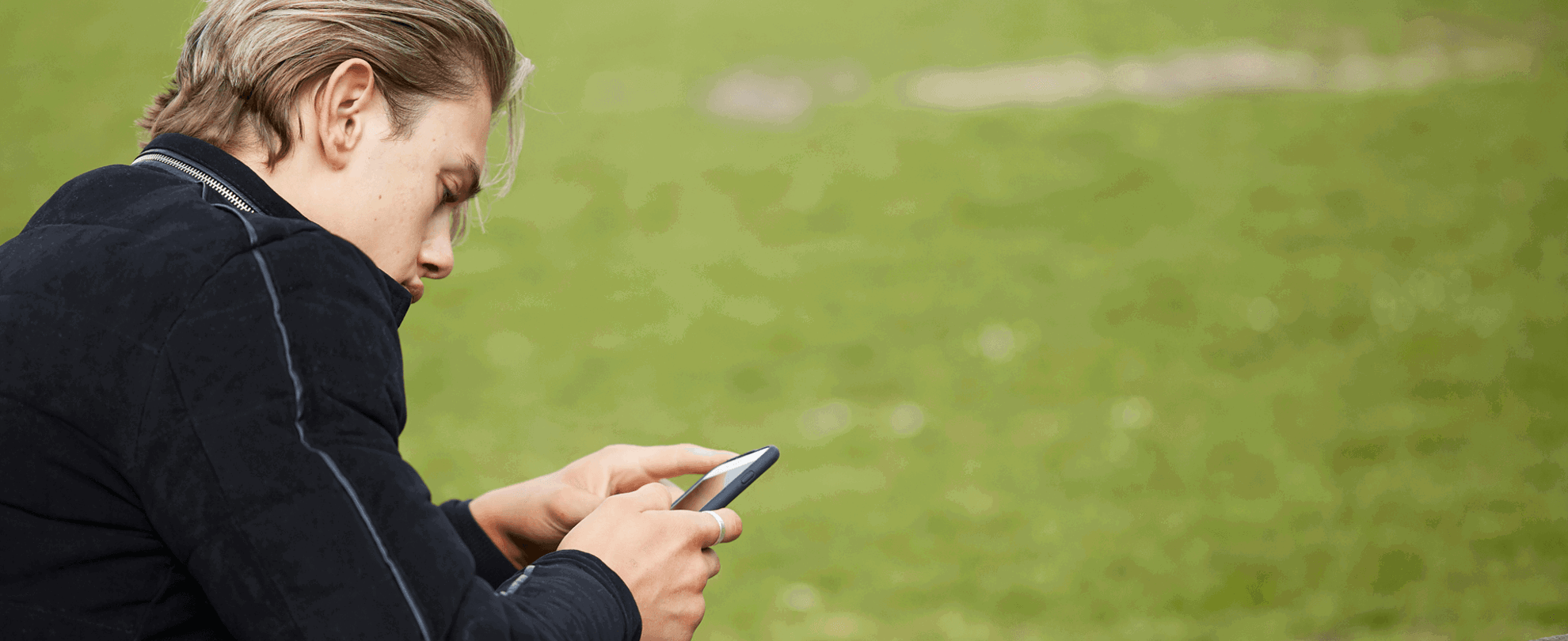side view close up of a boy wearing black jacket looking at his phone while sitting on a bench in a park