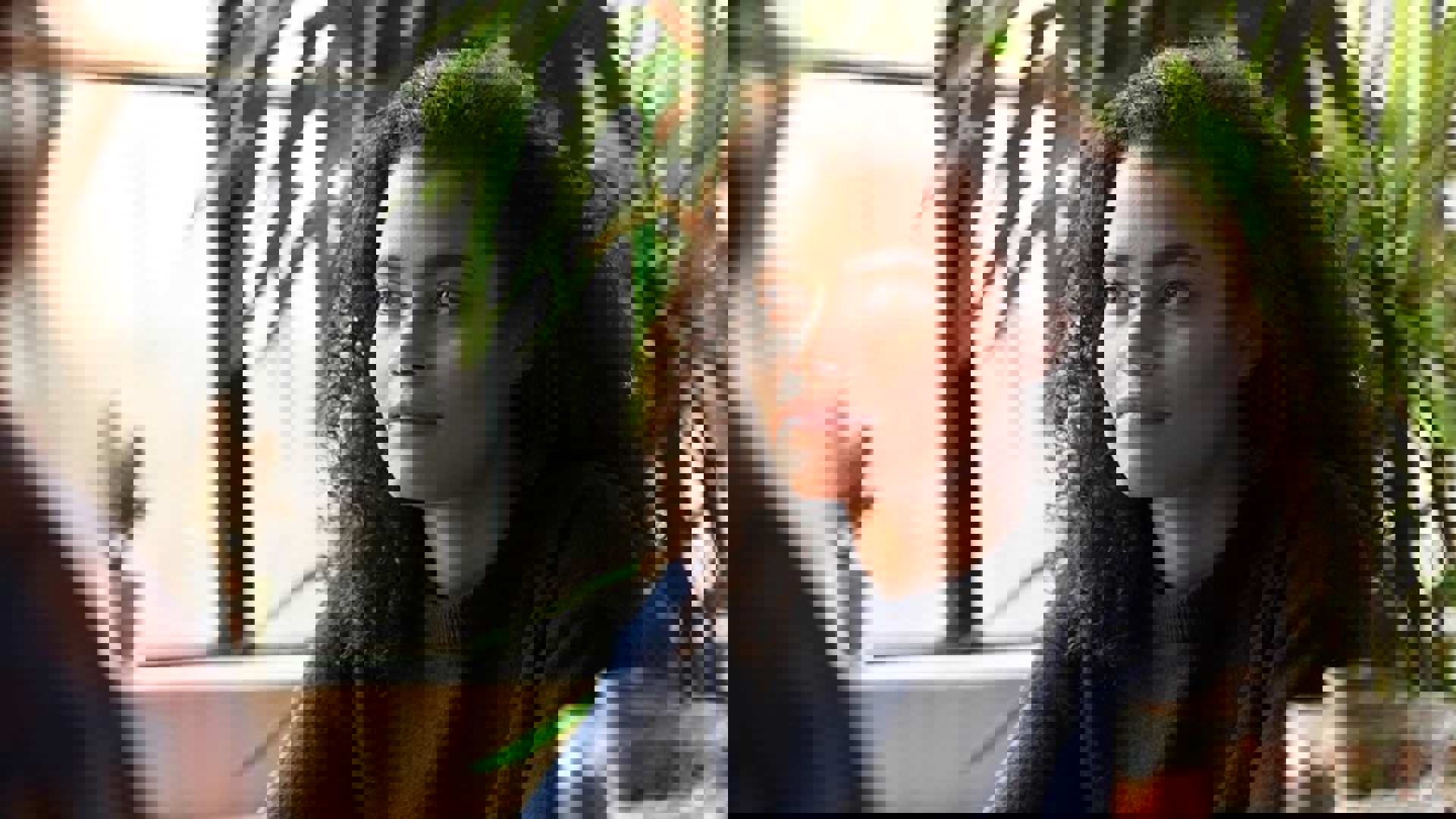 A girl with curly hair sitting in a cafe. She is looking away and smiling beside a window.