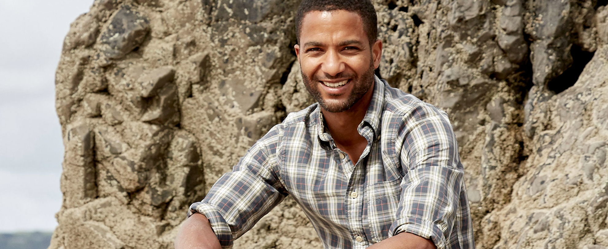 Sean Fletcher smiling and sitting on a rock by the coast
