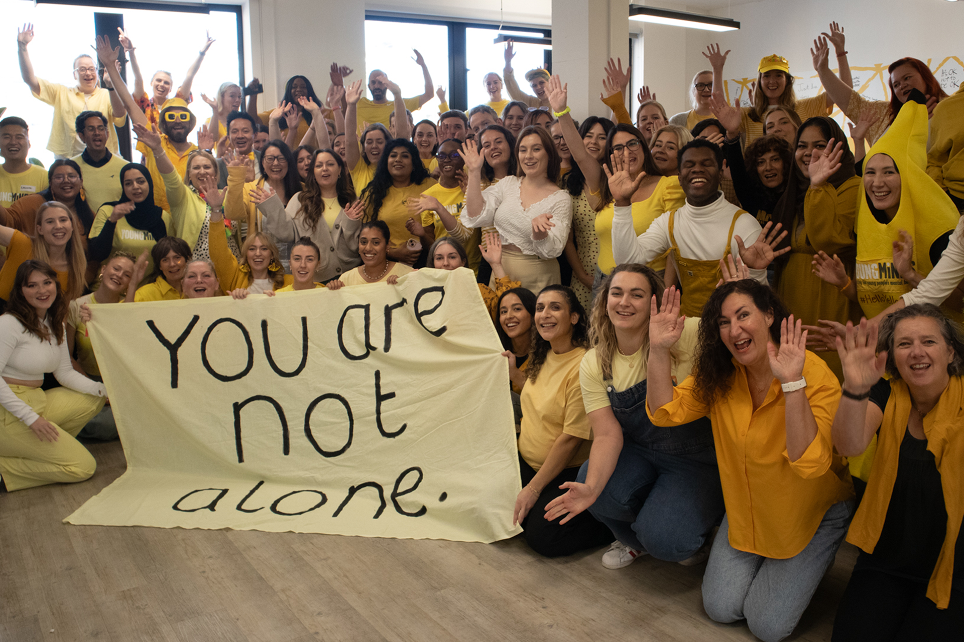 YoungMinds staff all wearing yellow for #HelloYellow 2023, posing with their hands in the air. They are holding a sign saying "You are not alone."