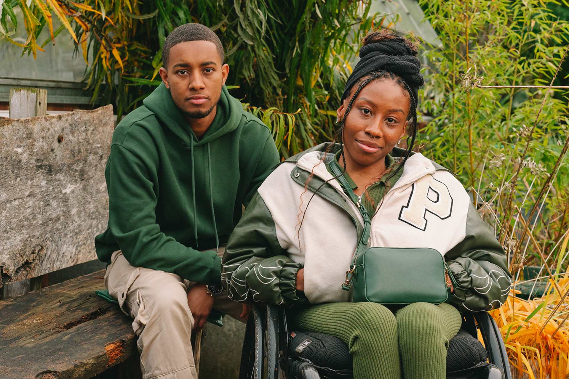 A young Black woman in a wheelchair and a young Black man on a bench, both staring at the camera looking serious.