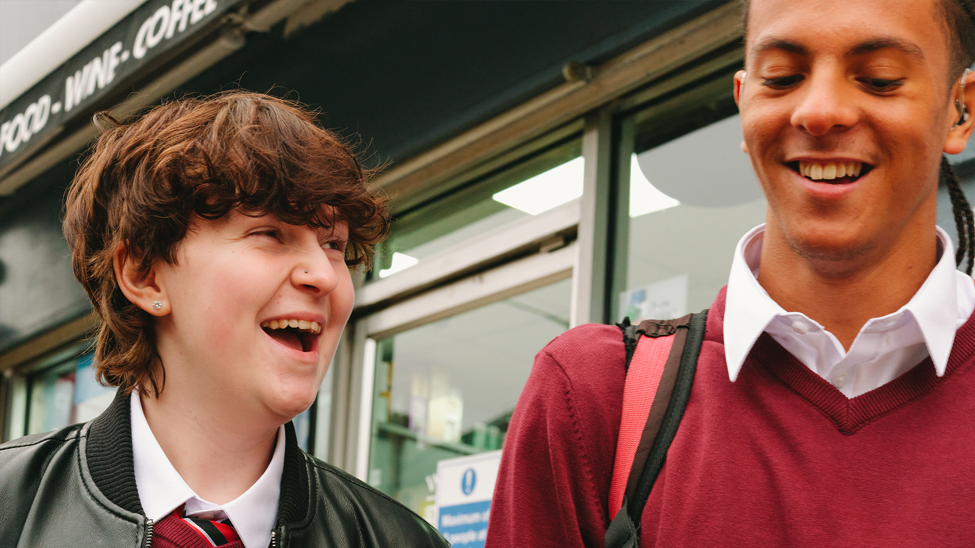 A Black teenage boy wearing a hearing aid laughing with a white non-binary teenager outside the shops.
