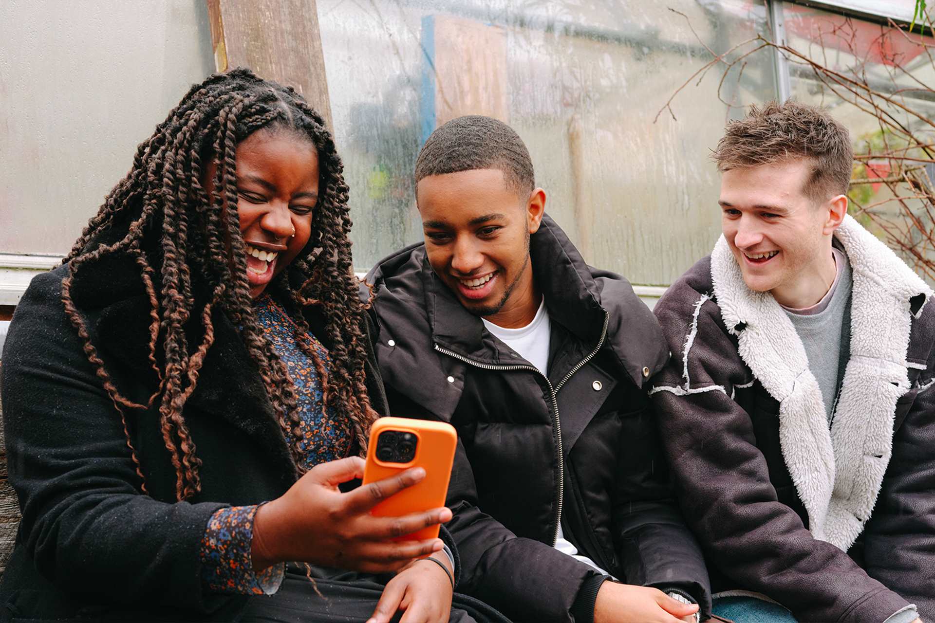 A young Black woman, young Black man and young white man, all sitting on a bench outside, looking at something on a phone and laughing.