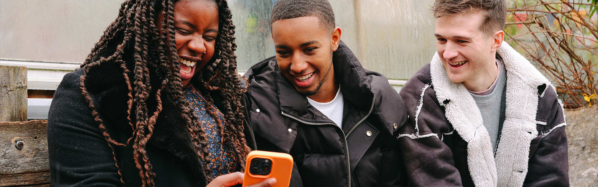 A young Black woman, young Black man and young white man, all sitting on a bench outside, looking at something on a phone and laughing.