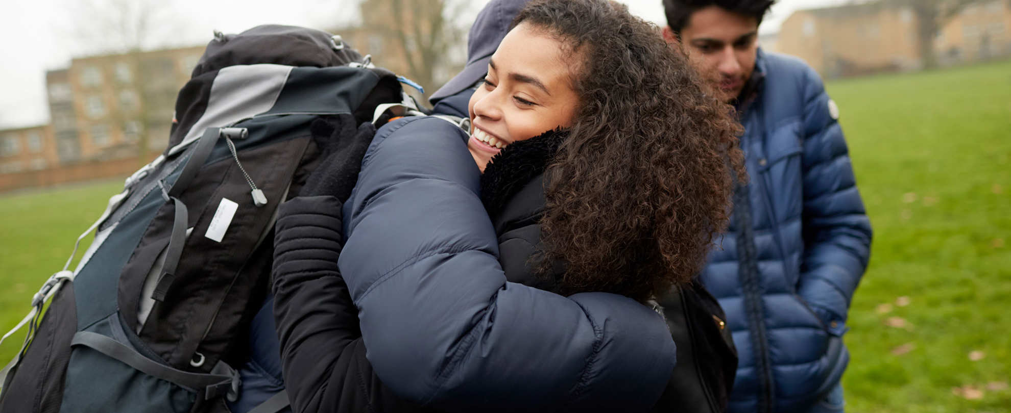 medium-shot-of-a-young-woman-with-curly-hair-smiling-while-hugging-another-person-with-face-unseen-and-wearing-a-cap-blue-jacket-and-a-huge-backpack-with-another-young-man-looking-down-with-school-building-on-background