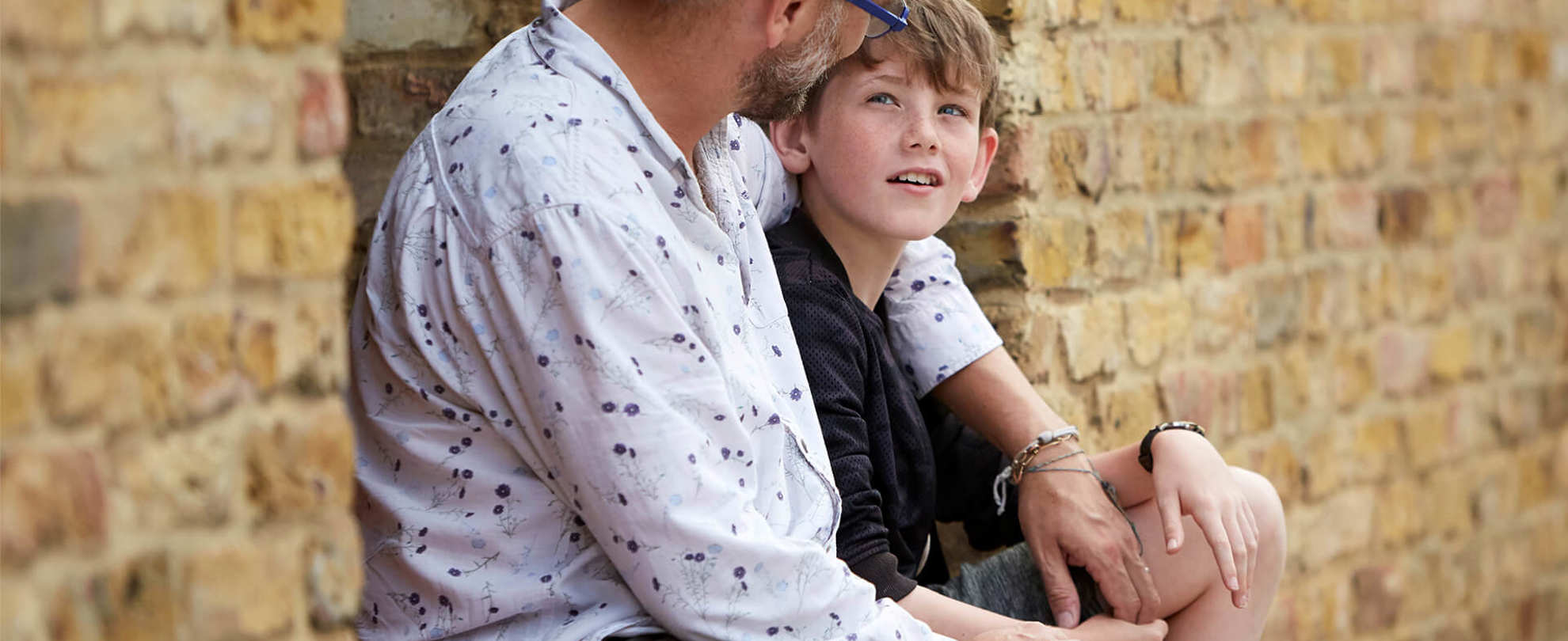 A father with his arm around his son sitting by a wall.