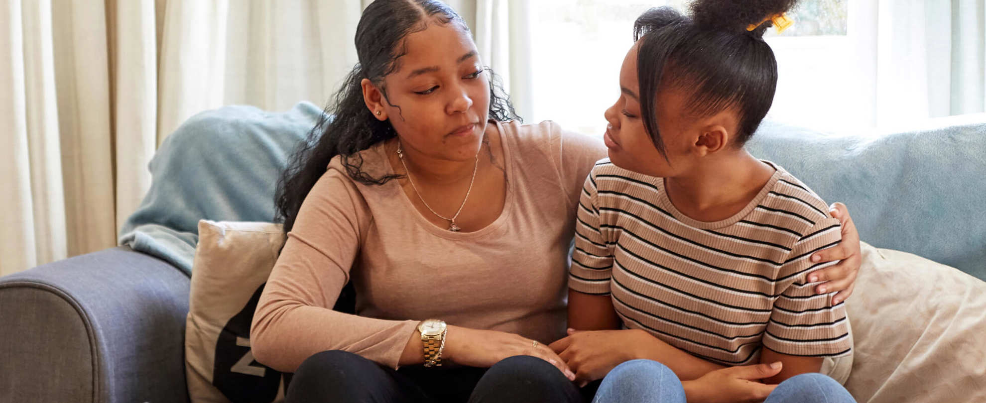 Two young people sit on a sofa with the person on the left putting an arm around the other. They both are looking at each other while talking.