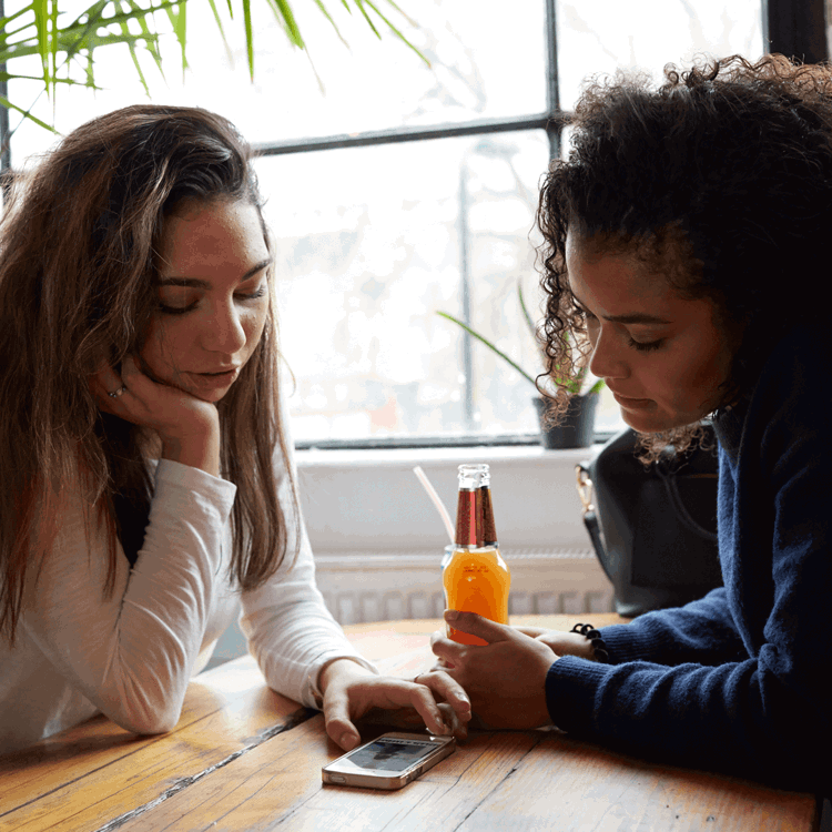 Two young women sit opposite each other at a wooden table in a restaurant. They are both looking at something on one of their smart phones, which is lying flat on the table between them,