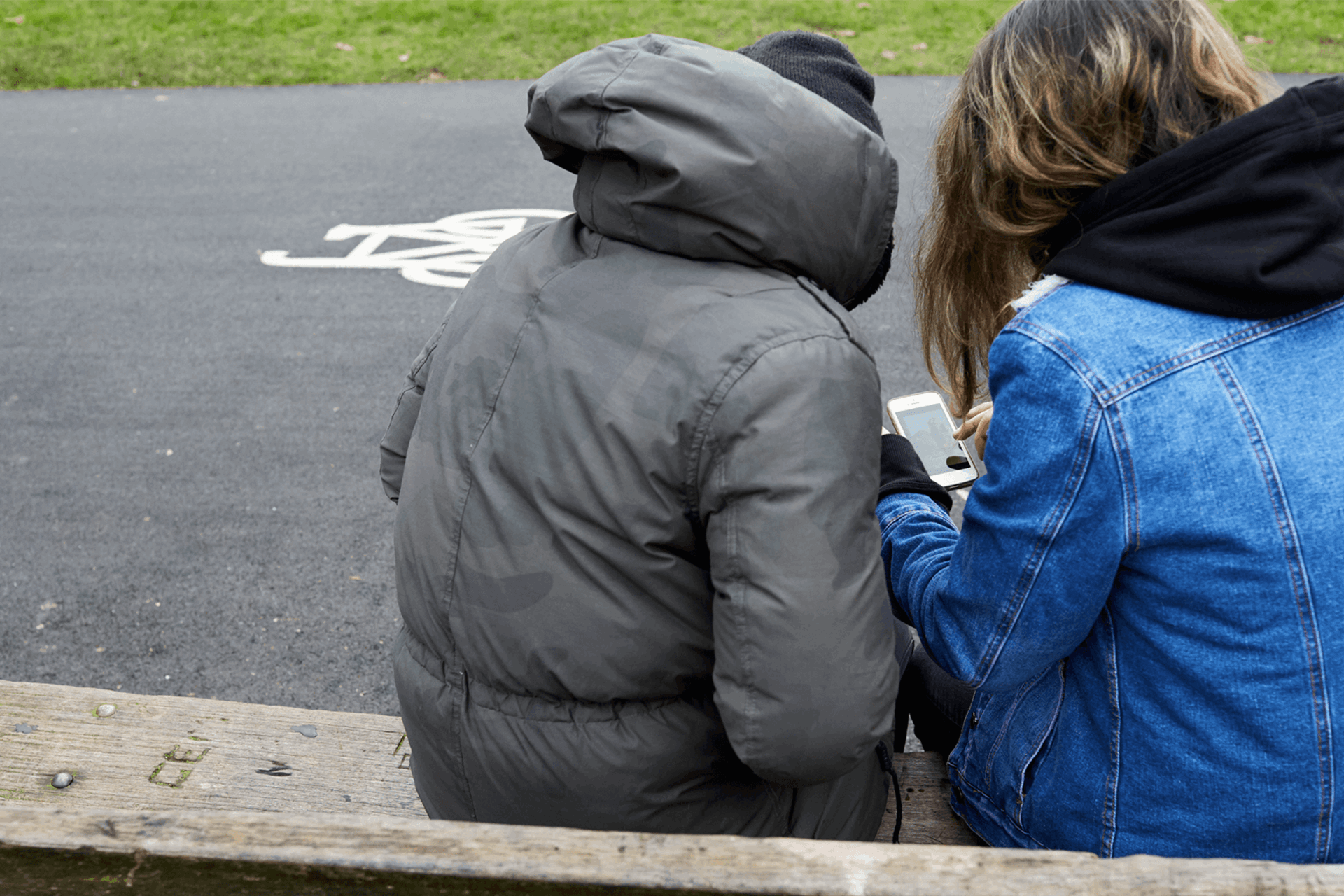 backshot of two young people wearing winter jackets with face unseen looking at their phone while sitting on a bench beside the street