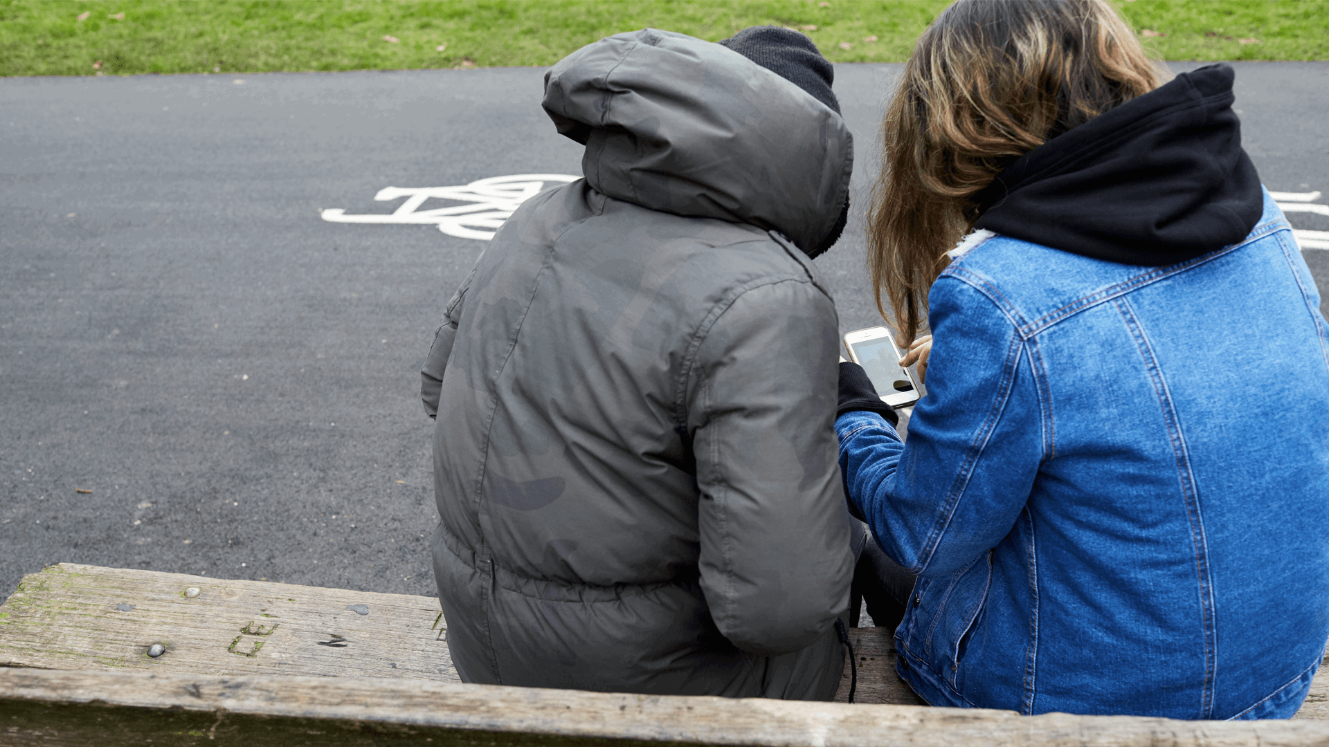 backshot of two young people wearing winter jackets with face unseen looking at their phone while sitting on a bench beside the street